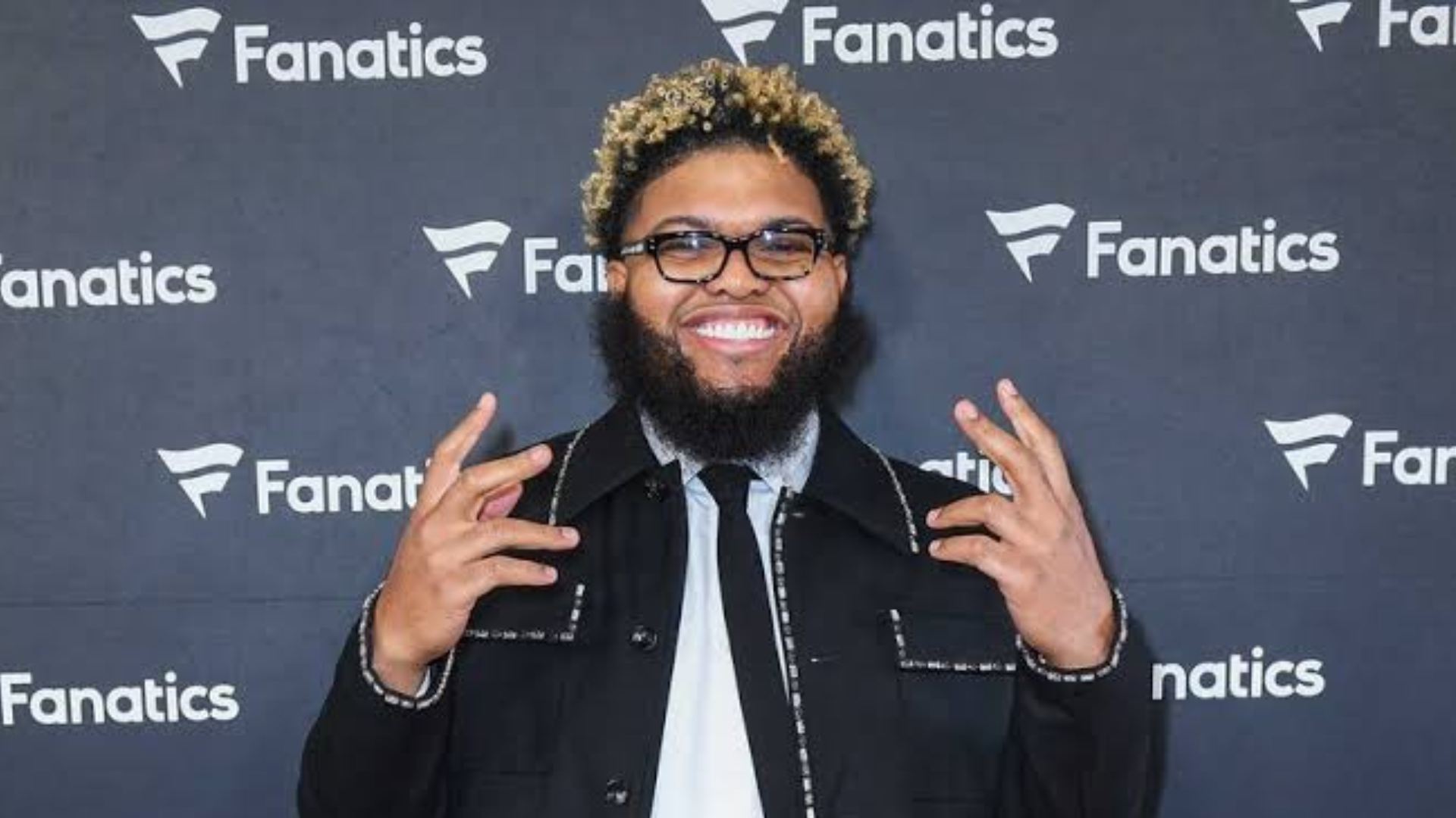 smiling druski with blond tipped curls black glasses and full beard posing with both hands raised before a fanatics backdrop