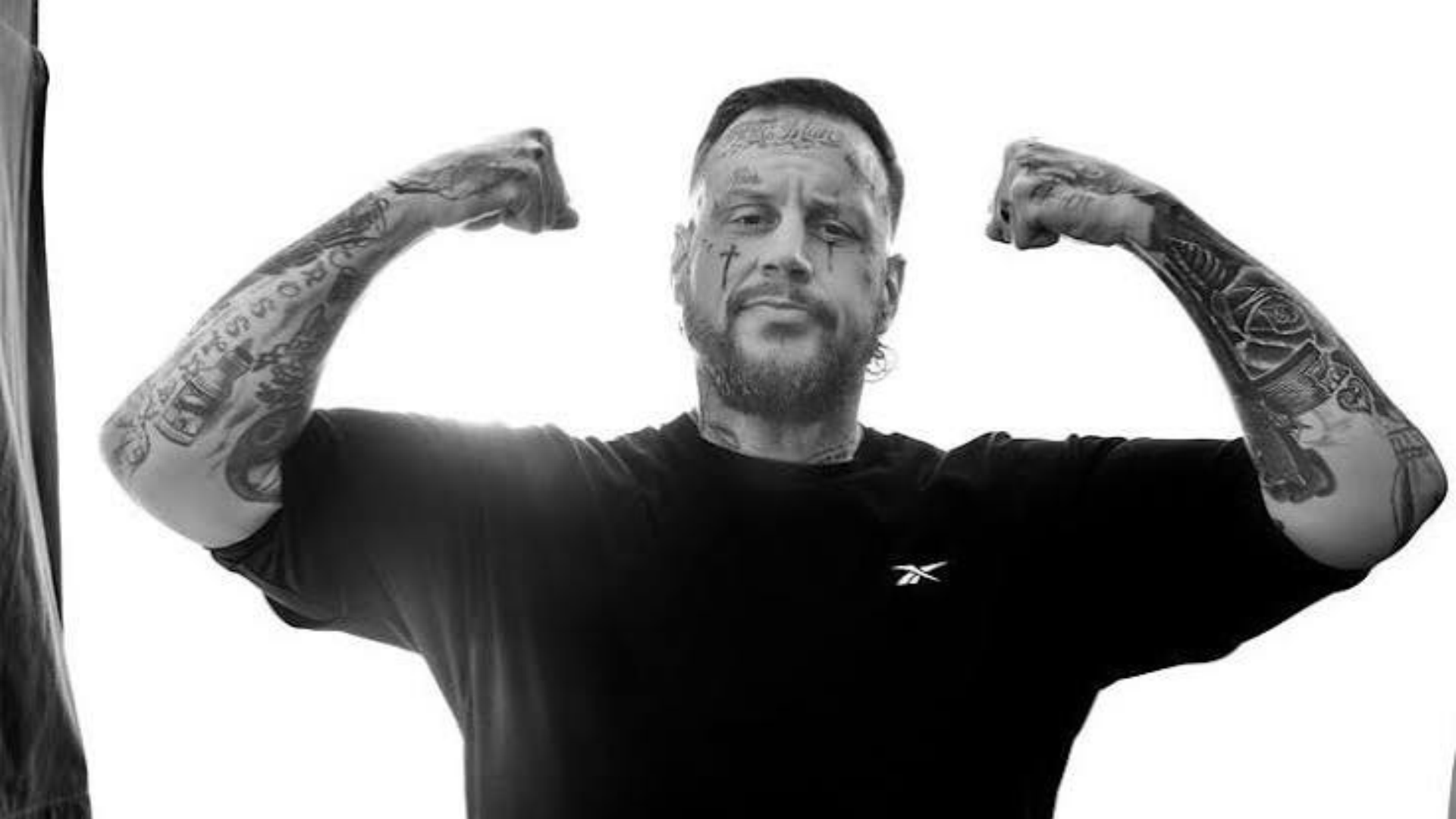 jelly rock in a black t-shirt flexing both arms in a high-contrast black-and-white portrait against a white background