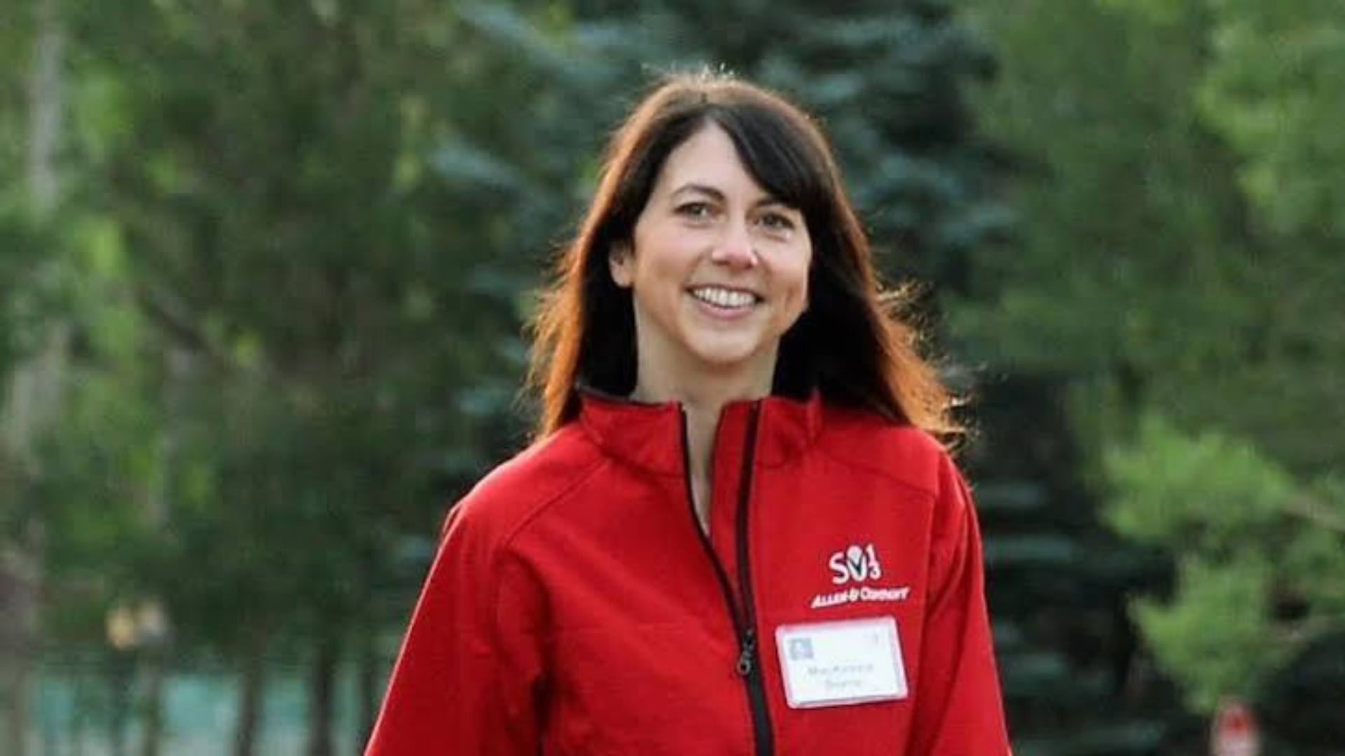smiling mackenzie scott in a bright red jacket standing outdoors against a blurred green tree and garden background in daylight