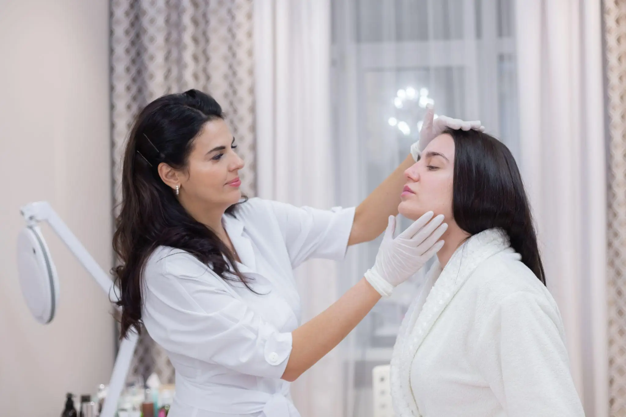 Esthetician examining woman's face in a spa clinic with patterned curtains in background