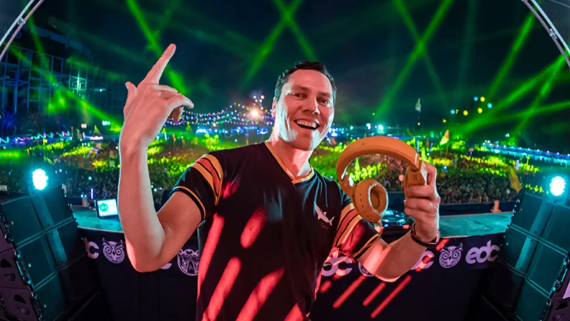 dj smiling on a festival stage while holding headphones, with green laser lights and a large crowd in the background