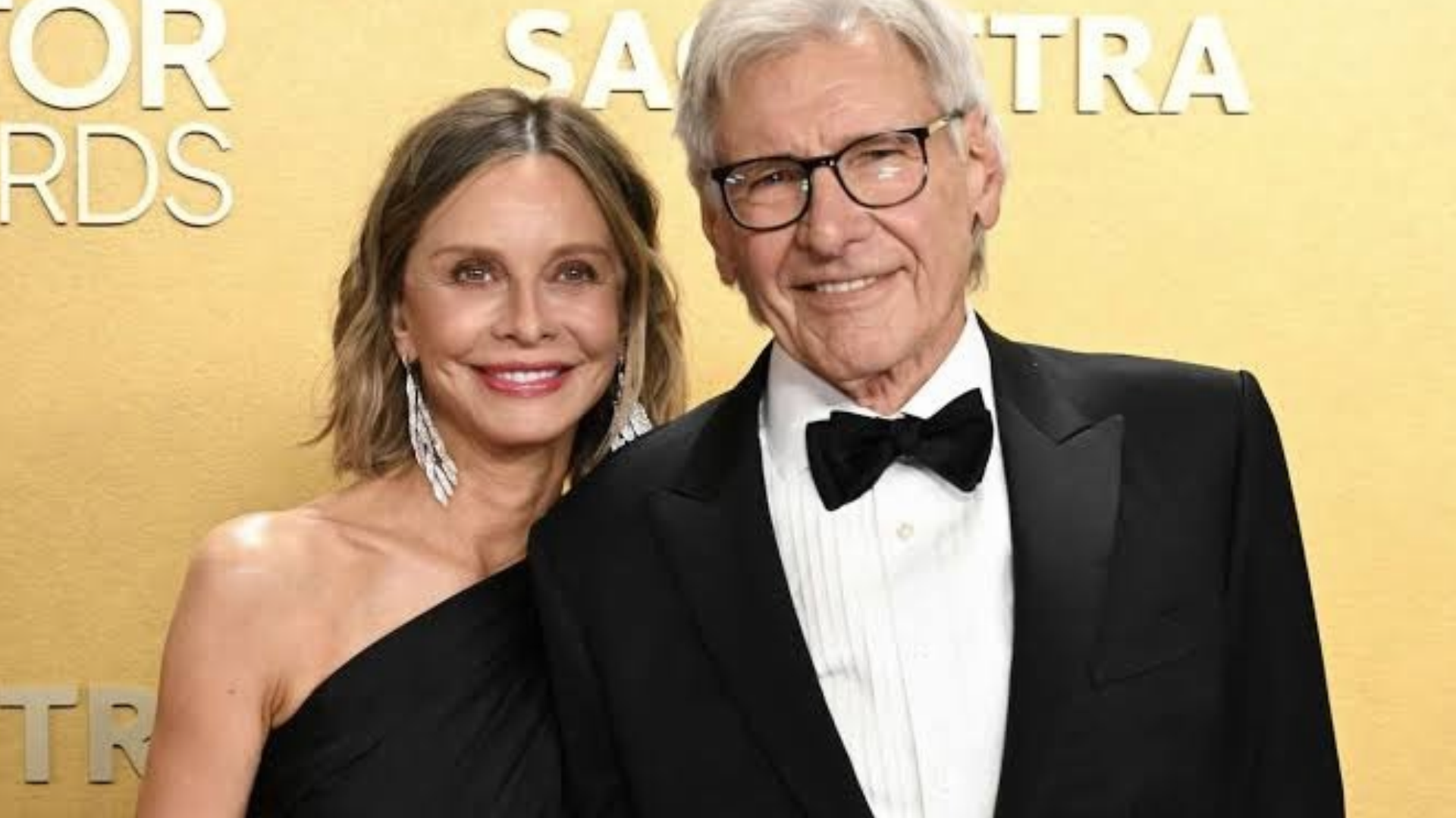 harrison ford in black tuxedo and glasses posing beside calista flockhart in black one shoulder gown against a gold sag awards backdrop