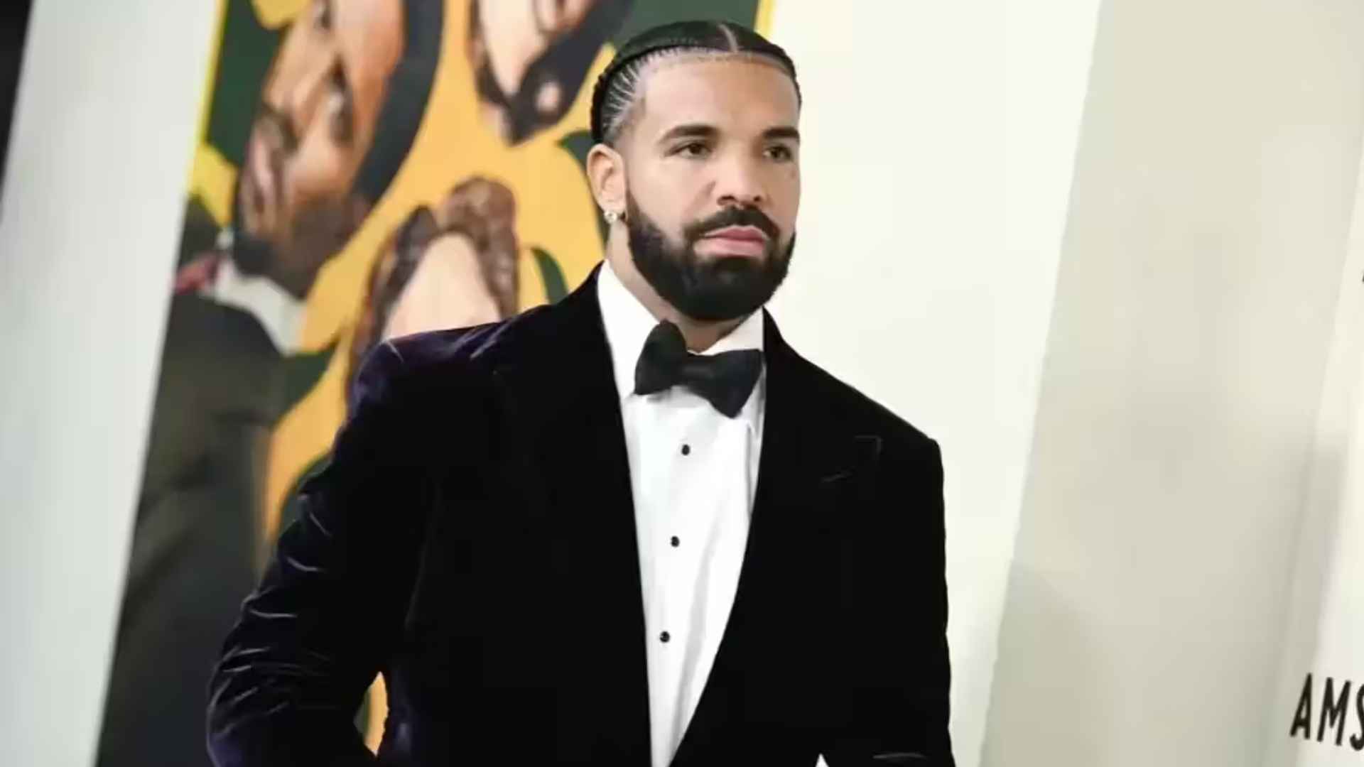 Drake wearing a black tuxedo with a bow tie while posing at a formal event with artwork in the background.