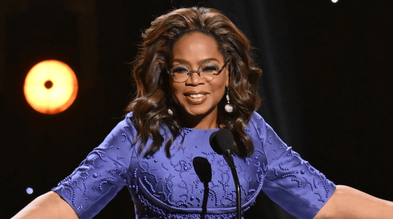 oprah winfrey speaks at a podium in a blue dress and glasses during a formal stage appearance