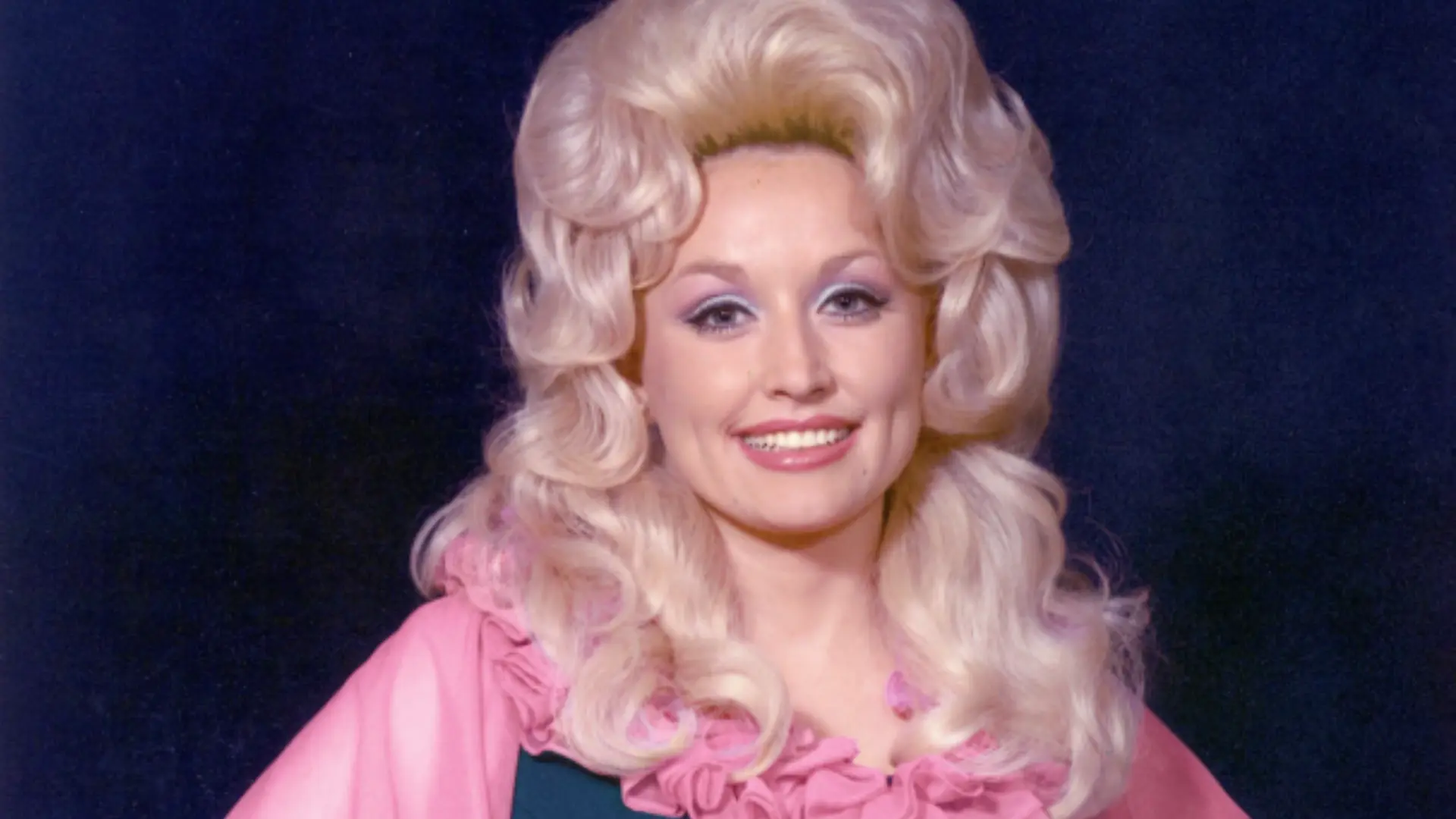 dolly parton in a vintage pink outfit with voluminous blonde hair, smiling in a close-up portrait against a dark backdrop
