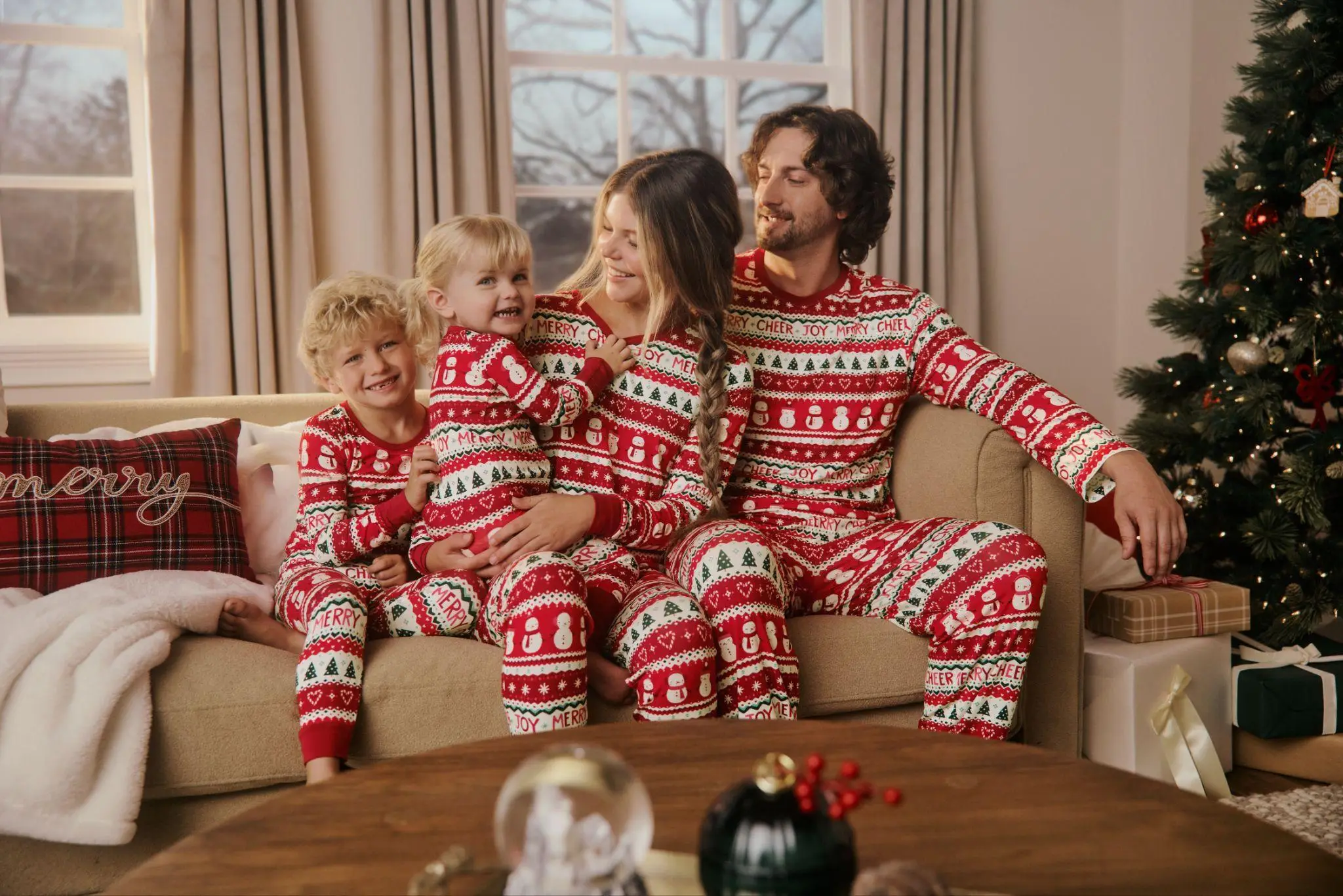 Family wearing matching Christmas pajamas on sofa next to decorated tree in cozy living room
