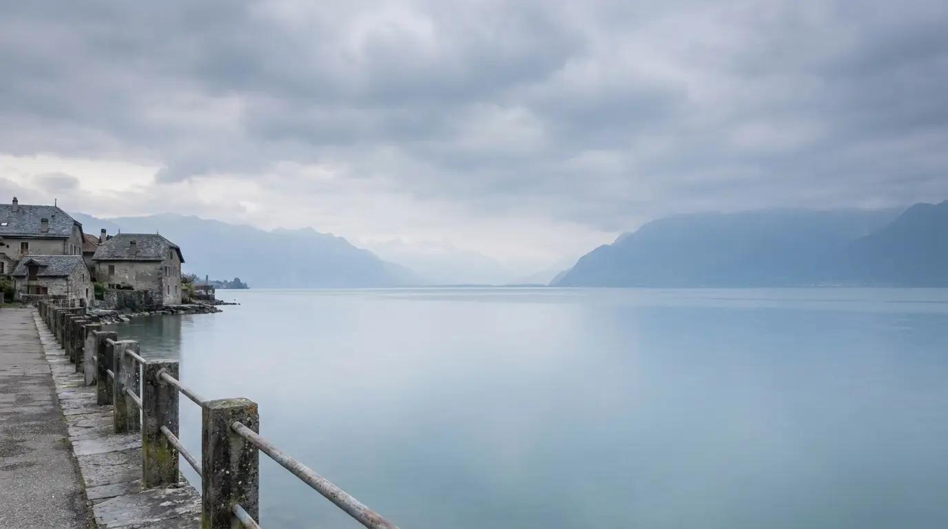 Stone buildings along a quiet lakeside promenade with overcast sky and distant mountains