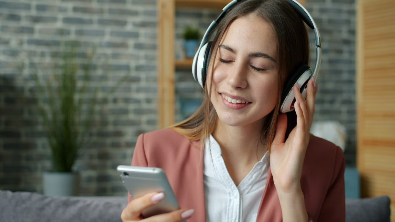 Person in pink blazer listening to music on headphones while holding a smartphone indoors